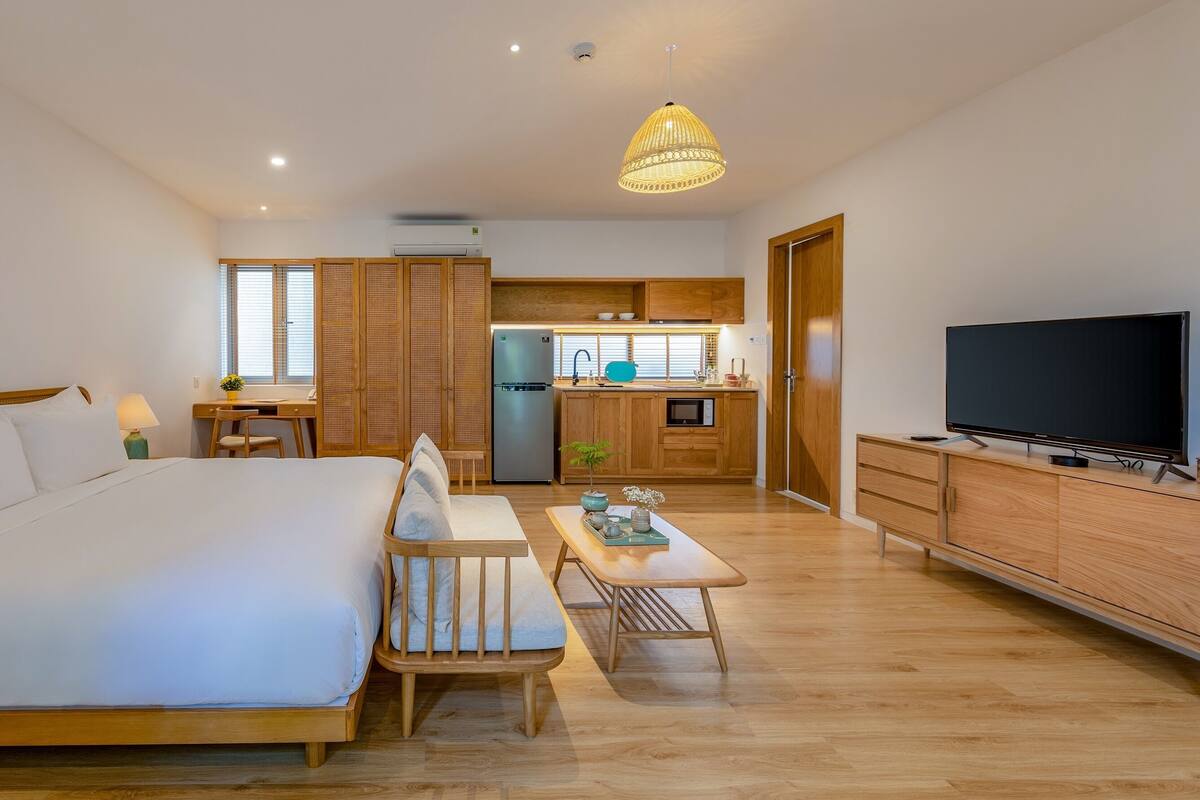 A modern hotel room with a bed, desk, kitchenette, and a TV.