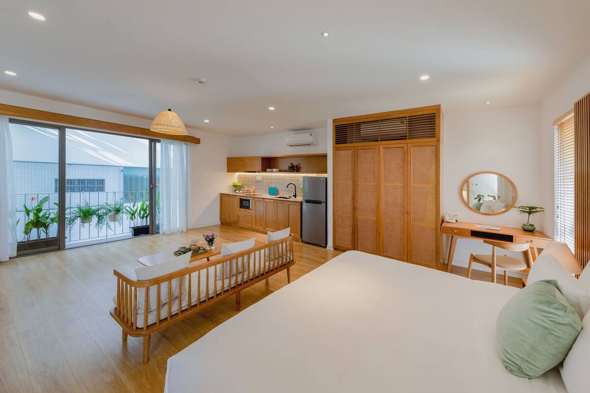 A modern hotel room with a large bed, a wooden desk, a kitchenette, and a balcony with plants.