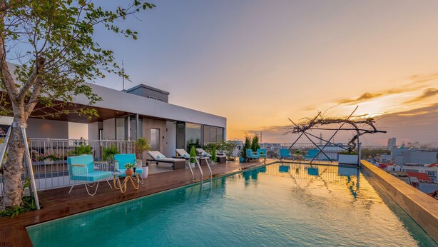 A rooftop pool area with lounge chairs, a wooden deck, and a view of the city skyline at sunset.