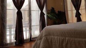 Ein Schlafzimmer mit einem Bett, großen Fenstern mit Vorhängen, Holzboden und einer Pflanze.