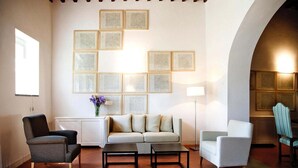 Ein Wohnzimmer mit Sofa, zwei Sesseln, einem Couchtisch und einer Heizung. An der Wand hängen Bilder in Rahmen, und auf der Heizung steht eine Vase mit Blumen.