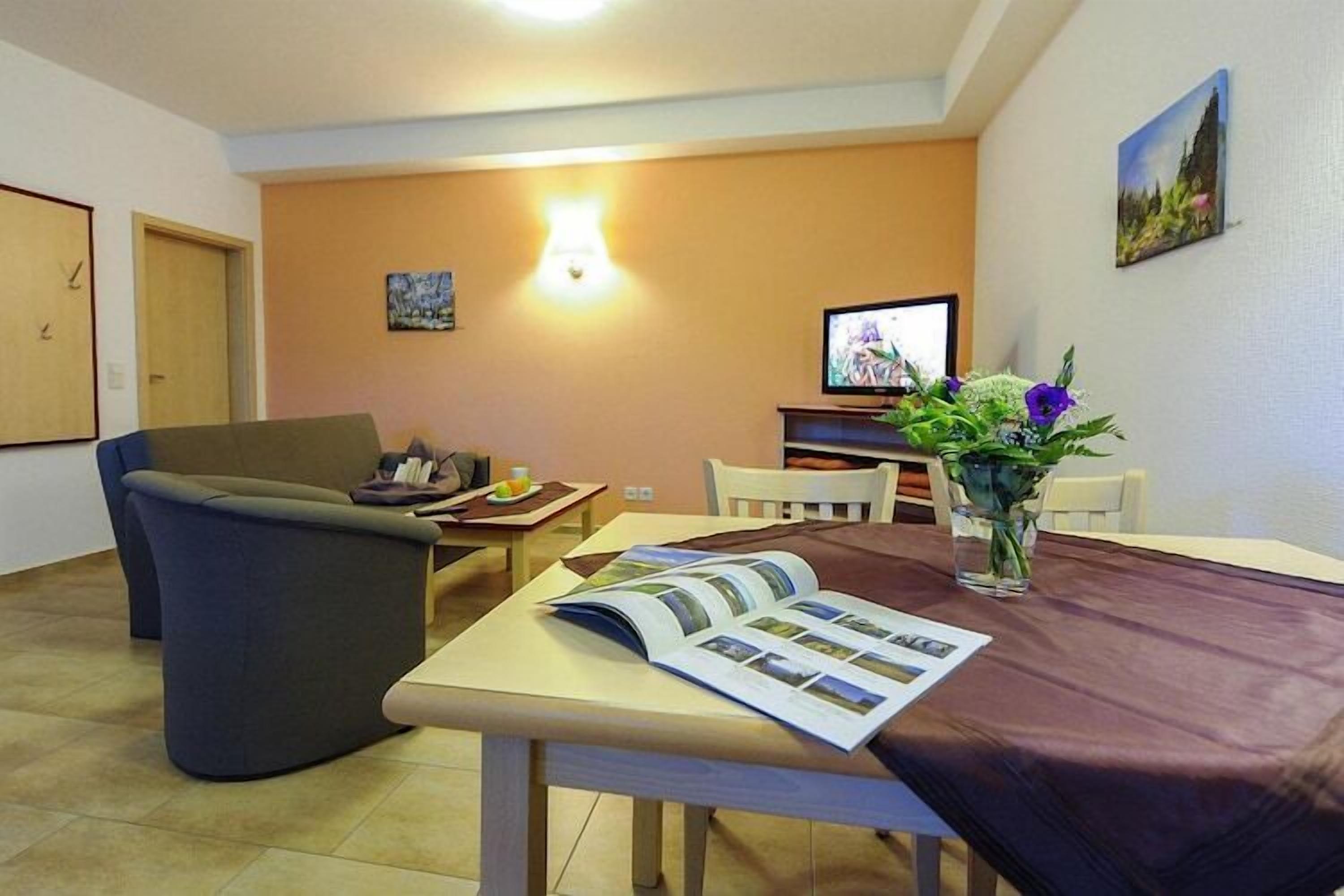 A cozy living room with a sofa, a dining table with a magazine, a TV, and a vase with flowers.