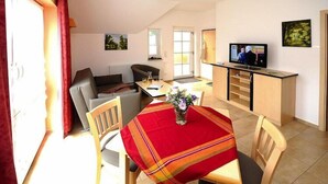 Ein gemütliches Wohnzimmer mit einer Couch, einem Esstisch mit roter Tischdecke und einem Fernseher auf einem Lowboard.