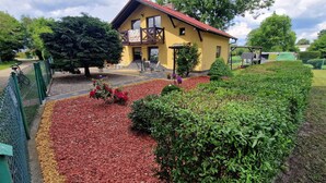 A yellow building with a red roof, a balcony, and a garden with red mulch and green plants.