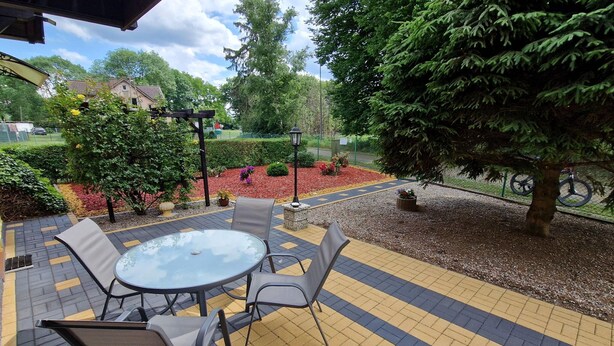 An outdoor patio with a round glass table and four chairs, surrounded by a patterned tile floor and various plants.