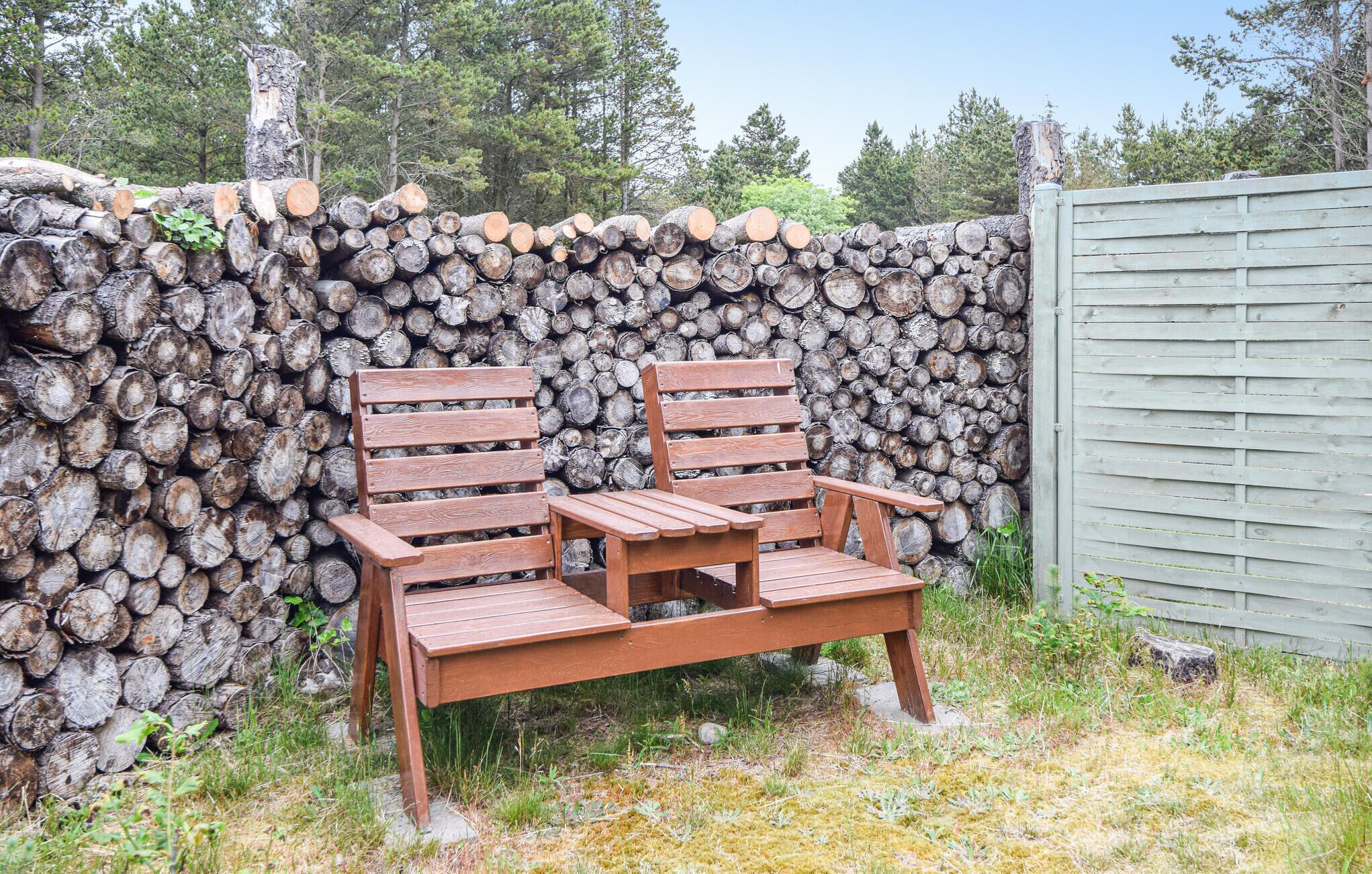 A wooden bench and chair set against a backdrop of stacked logs and a wooden fence.