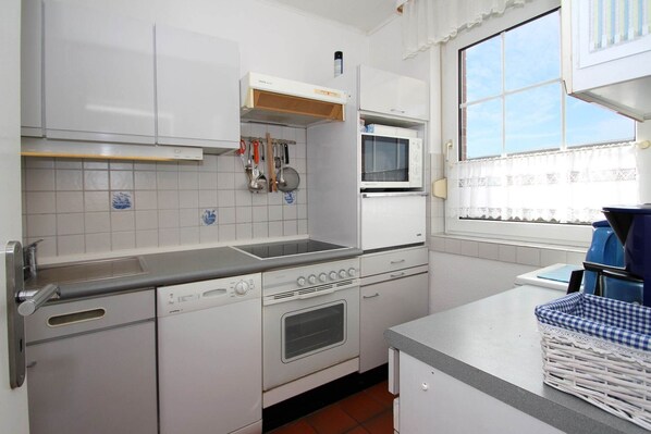 A compact kitchen with white cabinets, a built-in oven, a microwave, and a window with curtains. - Holiday Bungalow in Dornumersiel (Dornumersiel)