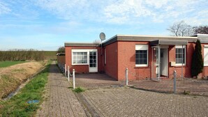 A red brick building with a satellite dish, a paved walkway, and a grassy field in the background. - Holiday Bungalow in Dornumersiel (Dornumersiel)