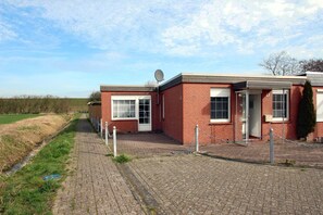 A red brick building with a satellite dish, a paved walkway, and a grassy field in the background. - Holiday Bungalow in Dornumersiel (Dornumersiel)