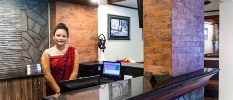 A reception desk with a digital screen, a sign indicating check-out times, and a reception area with a stone wall and a framed picture on the wall.
