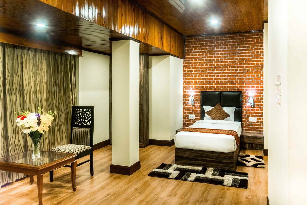 Hotelkamer met een bakstenen muur, een bed met hoofdeinde, een houten vloer, een glazen tafel met een bloemenvaas, en een stoel.