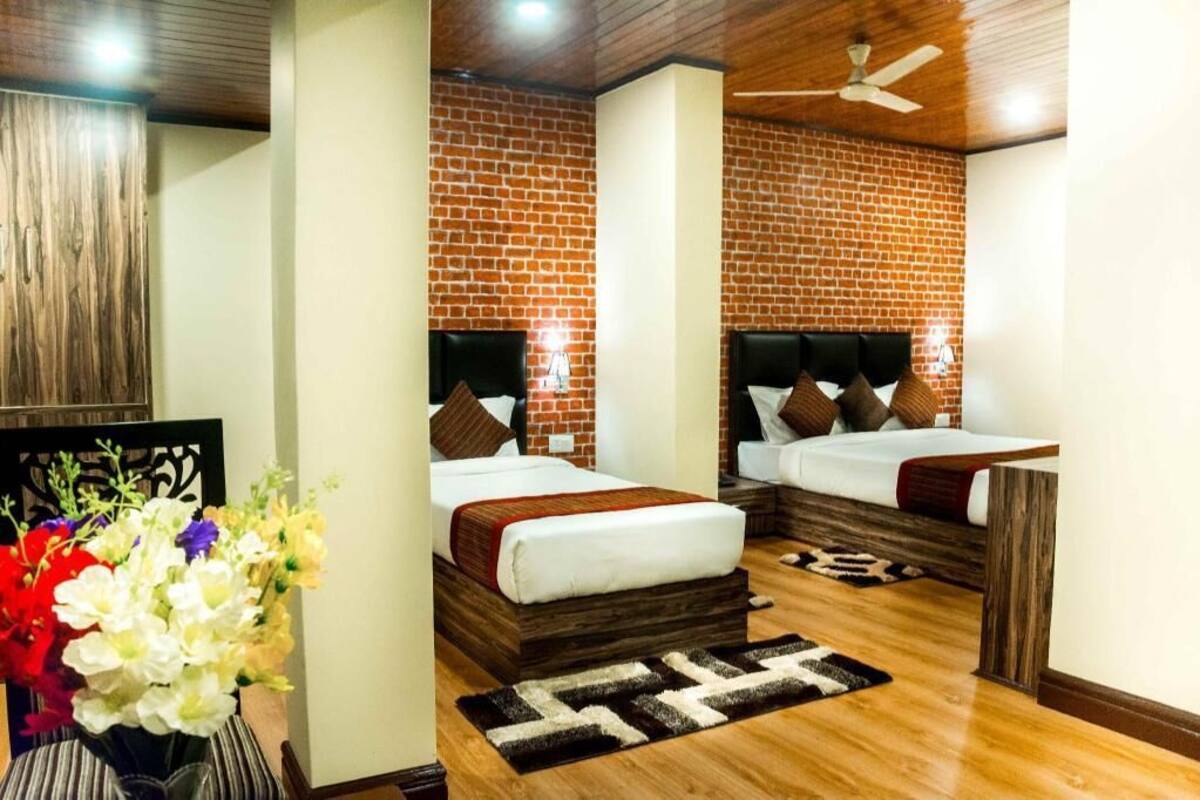 Een hotelkamer met twee bedden, een bakstenen muur, houten vloeren, een plafondventilator en een decoratieve vaas met bloemen.