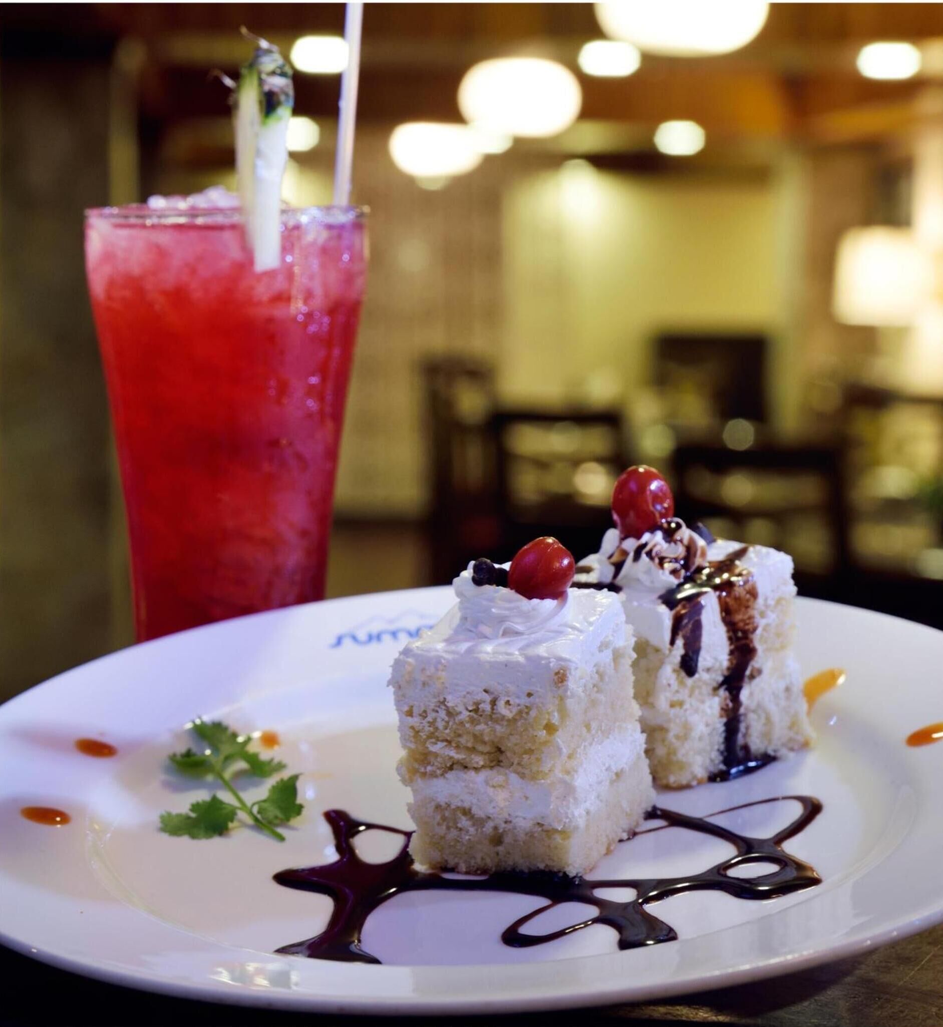 A dessert plate with two small cakes topped with whipped cream and cherries, and a glass of red beverage with a straw.