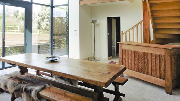 Une pièce spacieuse avec une grande table en bois et des bancs, un escalier et de grandes fenêtres donnant sur l’extérieur.