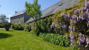 Un bâtiment en pierre doté d’un toit en pente, entouré d’une végétation luxuriante et de plantes grimpantes aux fleurs violettes.