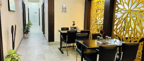 A hotel corridor with a dining area featuring a table set with chairs, a decorative wall, and potted plants.