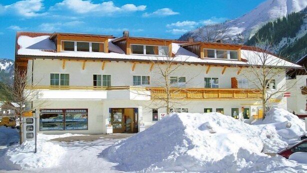 A multi-story building with a snow-covered roof, a visible storefront with signage, and a mountainous backdrop. - Apartment in Zugspitz Arena near Highline 179 (Bichlbach)