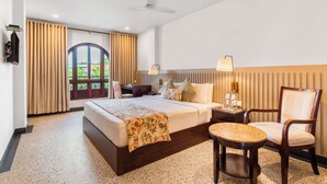 A hotel room with a bed, a chair, a desk, a lamp, a window with curtains, and a ceiling fan. - Hotel TSG Grand (Port Blair)