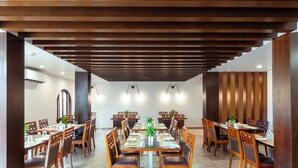 A spacious dining area with wooden tables and chairs, a central plant, and pendant lighting. - Hotel TSG Grand (Port Blair)
