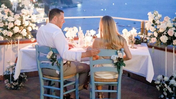 Una pareja cenando en una mesa al aire libre con vista al mar y un paisaje montañoso.