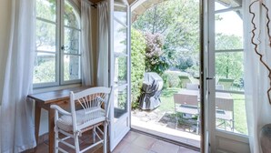 A room with a wooden table and a white chair, a glass door leading to an outdoor area with a barbecue and chairs.