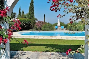 A swimming pool with a white pergola, surrounded by greenery and a clear sky.