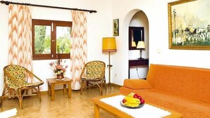 A room with a sofa, two chairs, a table with fruit, a lamp, and a painting.