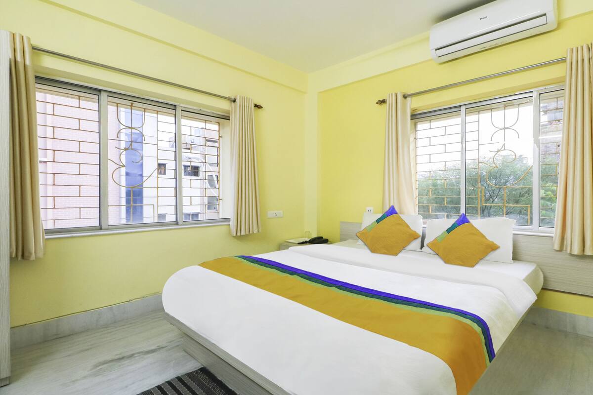 Une chambre d’hôtel avec un lit, deux fenêtres avec des rideaux, un climatiseur et une vue sur les bâtiments environnants.
