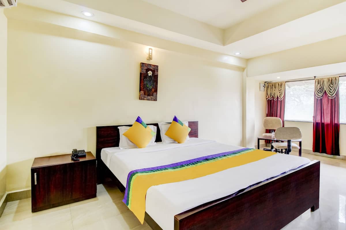 Quarto de hotel com uma cama grande, uma mesinha de cabeceira com telefone, uma cadeira, uma janela com cortinas e uma obra de arte na parede.