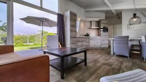 Een moderne woonkamer met een groot raam, een eethoek met rieten stoelen, een keuken met een inbouwoven en een grijze bank.