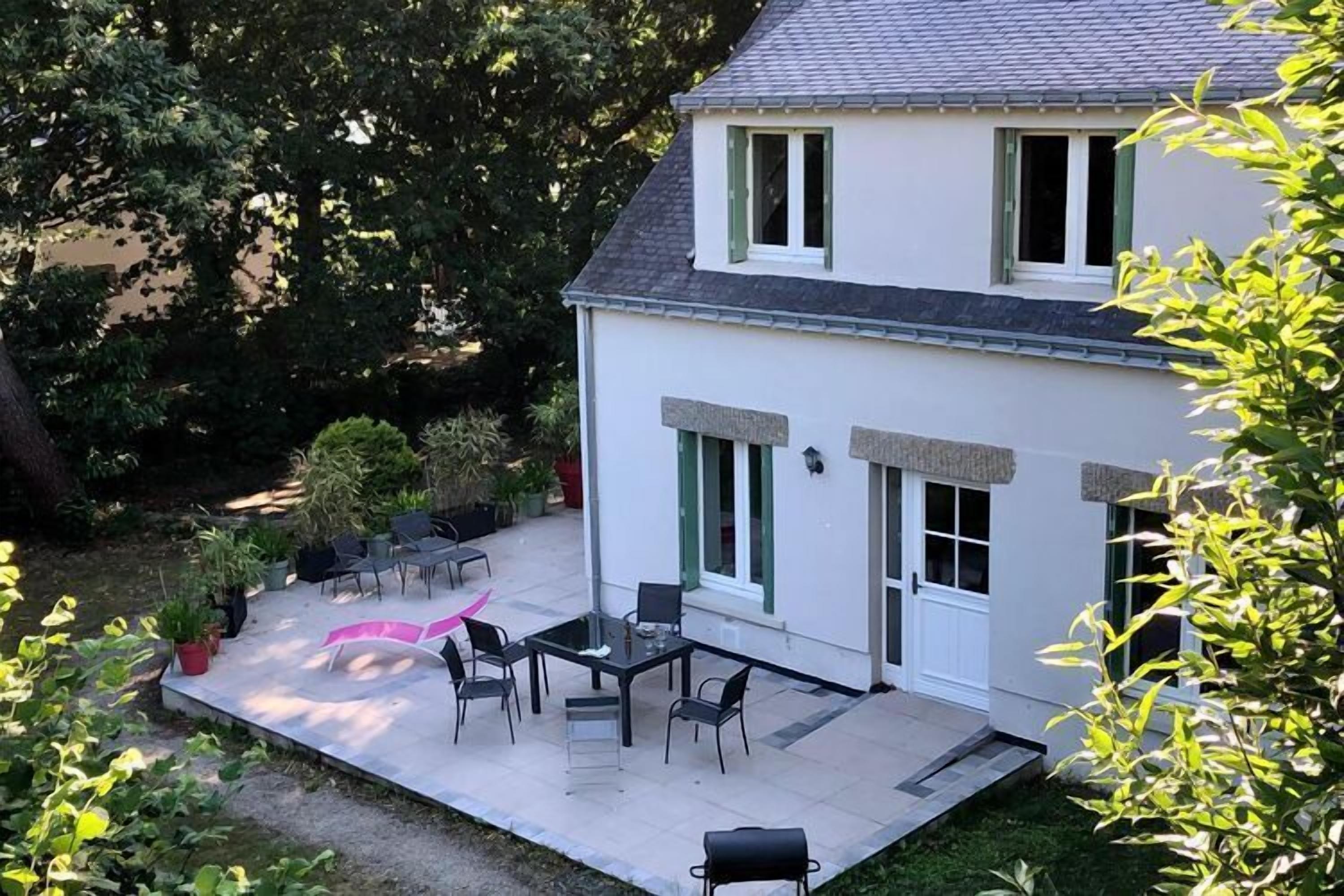 A two-story house with a patio area featuring a table and chairs, surrounded by greenery.