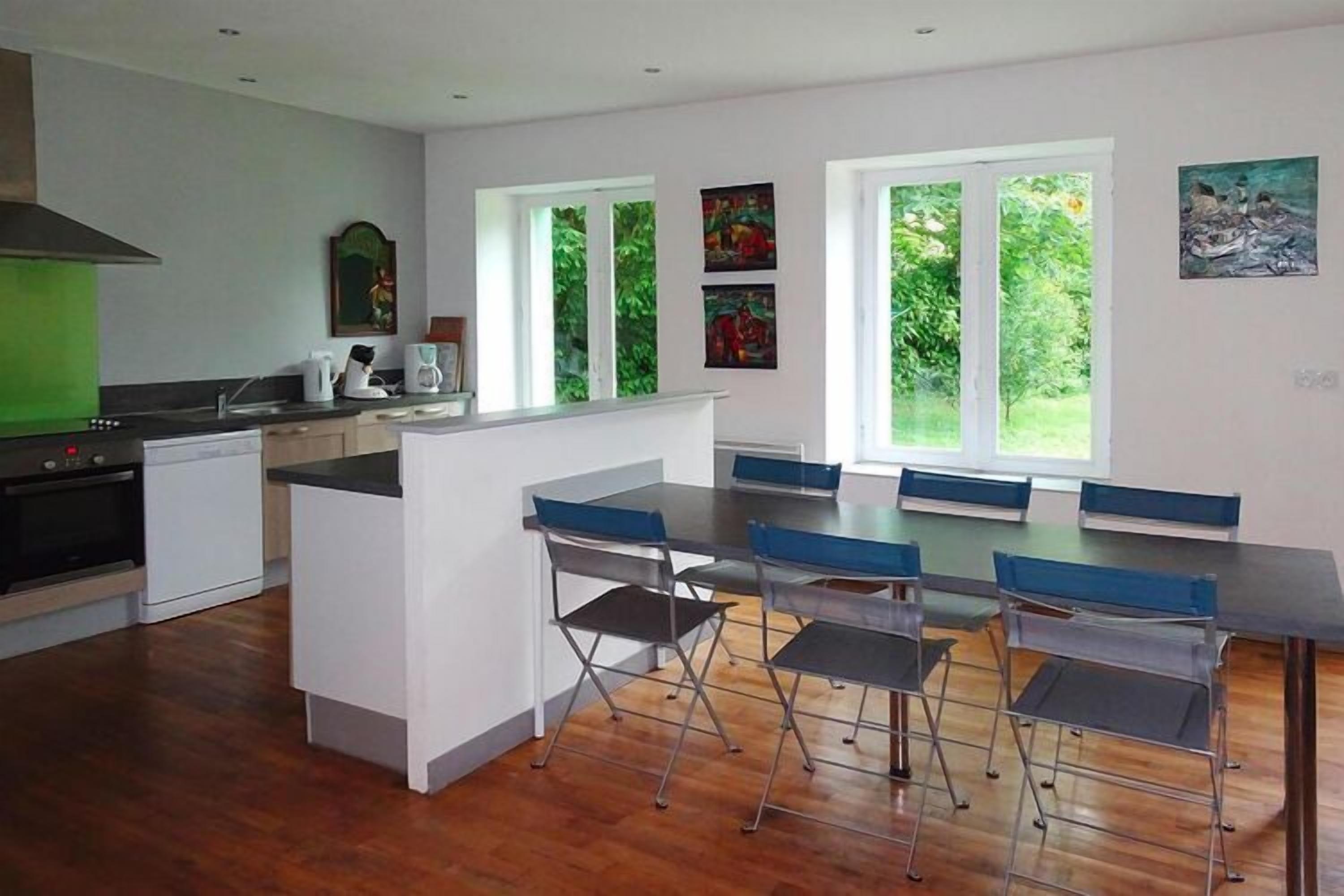 A modern kitchen with a central island, a dining area with foldable chairs, and large windows offering a view of greenery.
