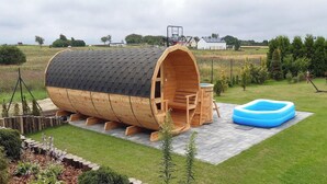 Uma estrutura em forma de barril de madeira com telhado, uma pequena área de estar e uma piscina inflável azul num quintal gramado.