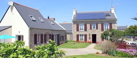 A two-story stone house with a sloped roof, a solar panel on the roof, and a well-maintained garden with flowers and shrubs.