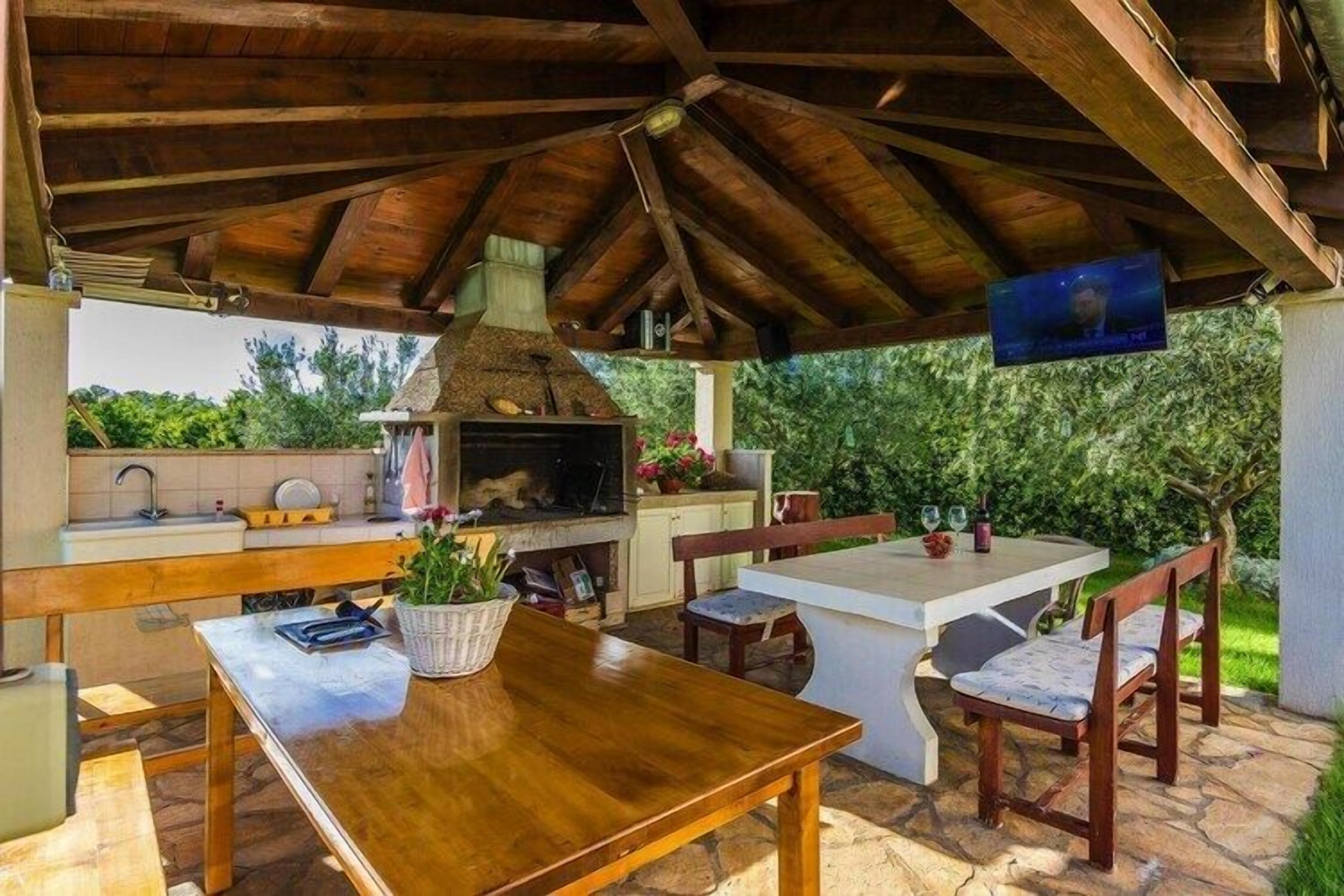Een buitenkeuken met een stenen aanrecht, houten tafel en stoelen, omgeven door groen.