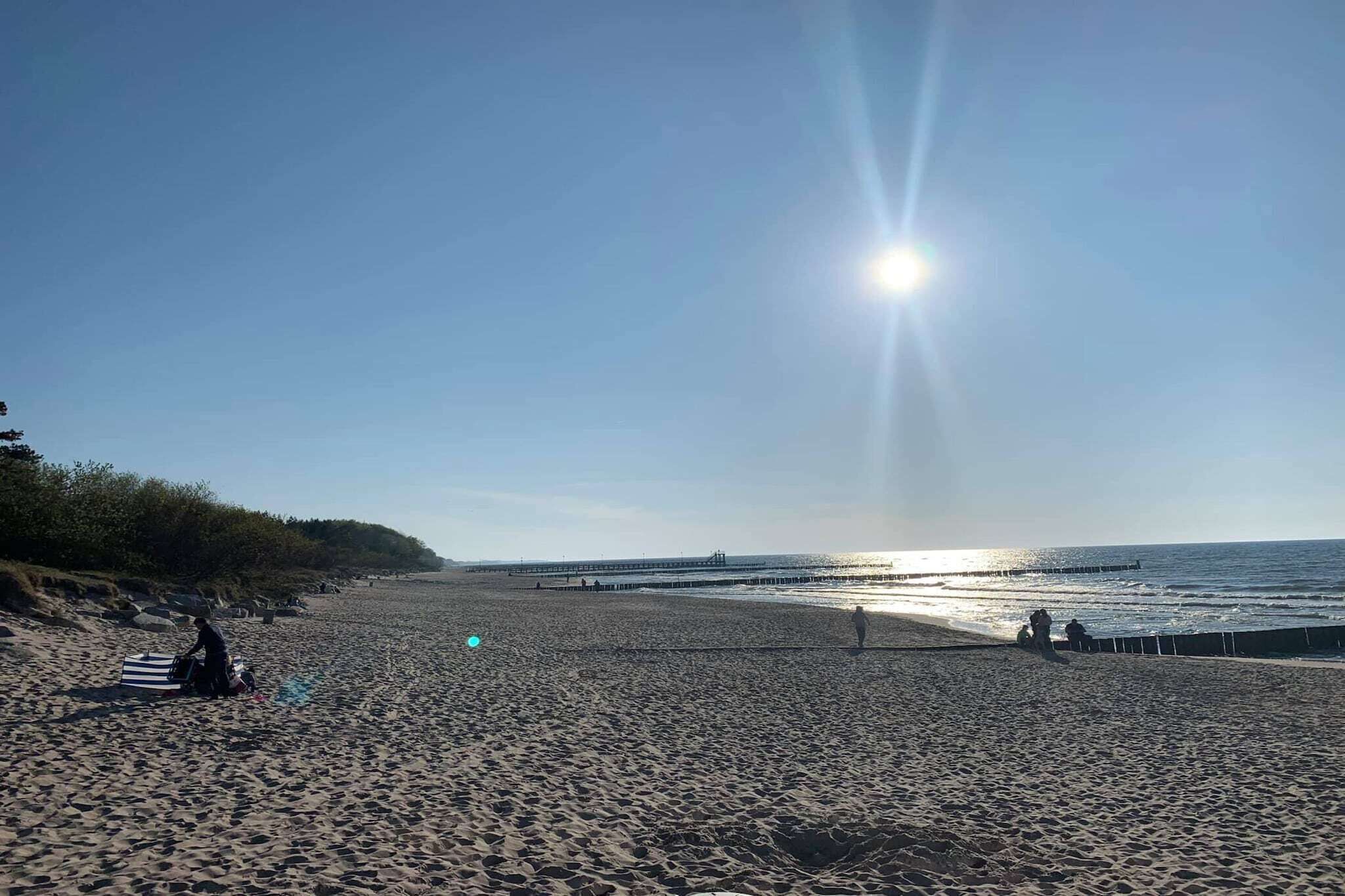 A beach with a pier extending into the water, clear skies, and a person sitting on a towel.