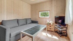 A cozy room with a grey sofa, a small table with a glass top, a TV on a wooden stand, and a window with a view of greenery. - Awesome home in Væggerløse with WiFi (Væggerløse)