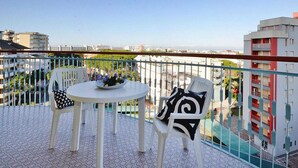 Un balcón con una mesa redonda y dos sillas, barandilla y vistas a edificios y vegetación.