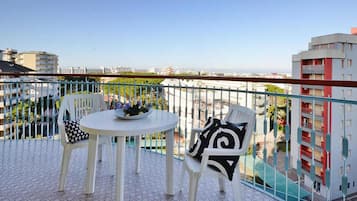 A balcony with a round table and two chairs, a railing, and a view of buildings and greenery.
