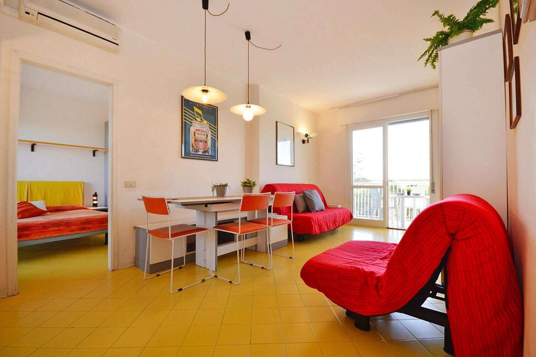 A compact living space with a bed, a dining table with chairs, a red sofa, and a balcony with a view.