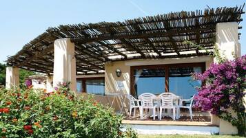 A covered outdoor seating area with white plastic chairs and a table, surrounded by lush greenery and vibrant flowers.