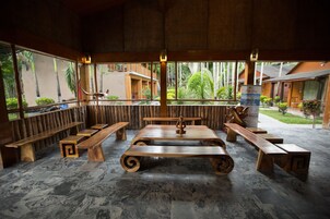 Un área de estar al aire libre con bancos y una mesa central, rodeada de amplios ventanales que ofrecen vistas a un jardín tropical y edificios.