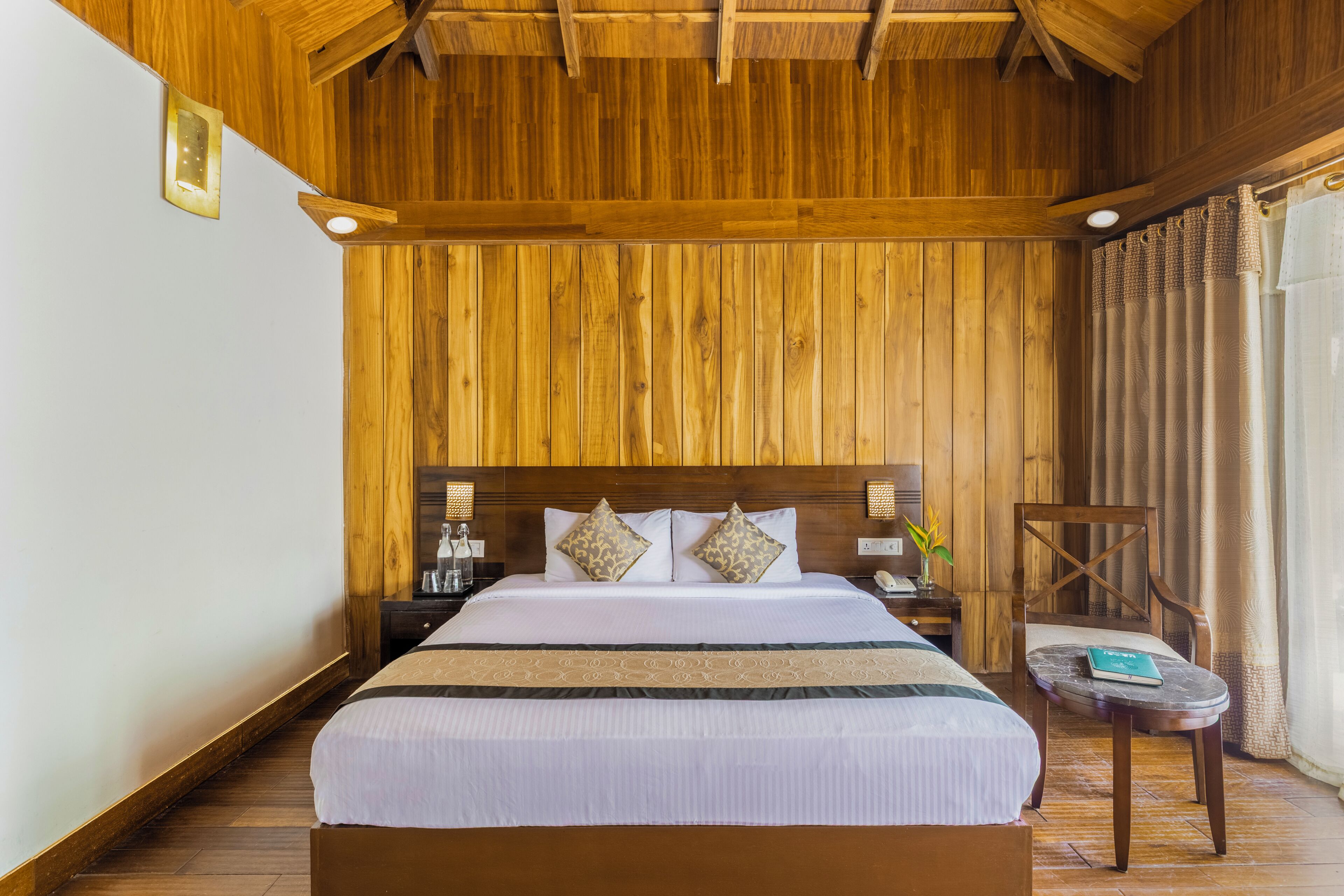 A hotel room with a wooden ceiling, a large bed with white bedding, two bedside tables, and a small round table with a chair.