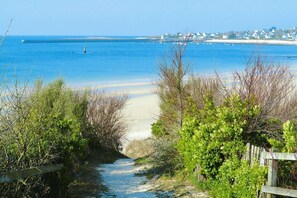 Uma trilha costeira leva a uma praia, com vista clara para o mar e uma linha de costa distante com casas.