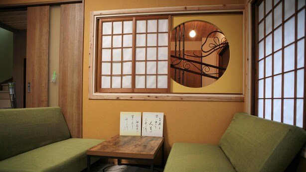 A room with a wooden table, two green sofas, a window with a decorative metal design, and two signs with Japanese text. - Maru House - Hostel (Osaka)
