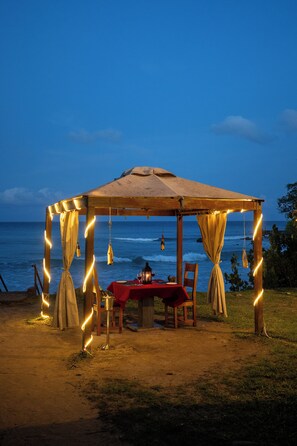 A beachside dining setup with a table under a canopy, surrounded by chairs and a wooden structure. - TSG Aura (Port Blair)
