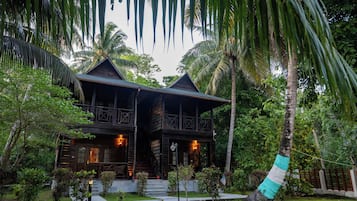 A wooden house with a balcony, surrounded by lush greenery and palm trees.