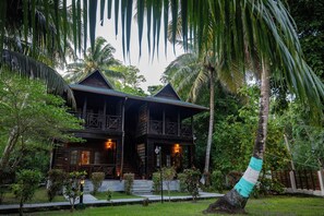 A wooden house with a balcony, surrounded by lush greenery and palm trees. - TSG Aura (Port Blair)