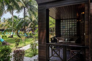 Una estructura de madera con balcón que da a un jardín con palmeras y una zona de piscina.