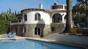 A poolside view of a building with a stone facade, a pool, and blue lounge chairs.